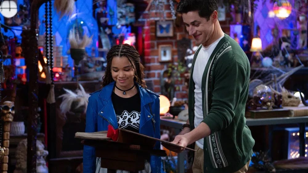 An adult Justin Russo stands smiling in a green patterned cardigan over a white t-shirt and khaki pants alongside Billie, a young wizard with braided hair wearing a bright blue leather jacket over a black graphic t-shirt and a plaid skirt, as they look through a large ancient spellbook resting on a wooden podium inside a cluttered, magical room filled with eclectic artifacts, glowing orbs, and brick walls. An older man with dark hair, wearing a white t-shirt and an open green cardigan over tan pants, stands next to a young girl with braided hair wearing a bright blue leather jacket and black t-shirt over a plaid skirt as they both look down smiling at a large open book resting on a wooden podium in a cluttered room filled with miscellaneous trinkets, books, and lights.