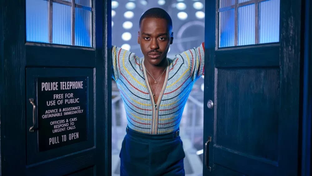 The image shows Ncuti Gatwa as the Fifteenth Doctor in the sci-fi television series Doctor Who. He is standing in the doorway of the TARDIS, the Doctor's time-traveling craft that resembles a blue British police box.The Doctor is wearing a short-sleeved, zip-up knit shirt with horizontal multicolored stripes (red, yellow, blue, and white) on a light blue base, paired with dark blue trousers. He has a serious expression, dark hair, and a mustache. Behind him, the interior of the TARDIS is visible with glowing blue and white circular lights, while the front of the police box features a sign that reads "POLICE TELEPHONE / FREE FOR USE OF PUBLIC".This specific shot was frequently used in promotional materials for the 2024 season of the show. Gatwa portrayed the Doctor for two seasons before the character was depicted as regenerating in the season finale.