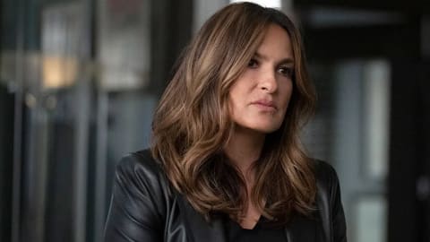 An image showcases American actress Mariska Hargitay, widely recognized for her iconic role as Captain Olivia Benson on the long-running NBC drama series Law & Order: Special Victims Unit, posing in a modern indoor setting with a serious, contemplative expression. Her mid-length, warm brunette hair is styled in voluminous, soft waves with subtle caramel highlights that frame her face elegantly. She wears a sophisticated ensemble featuring a sleek, tailored black leather blazer layered over a simple, matching black top, projecting a sharp, authoritative, and professional aesthetic. The background is softly blurred, hinting at an office or studio environment with vertical architectural elements and muted lighting that keeps the visual focus entirely on her compelling presence.