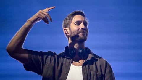 An outdoor musical performance features a man, captured from the chest up, with a short beard and mustache. He is wearing a dark, unbuttoned short-sleeved shirt over a white undershirt, with black headphones resting around his neck. His right arm is raised with his fingers pointing upward, and he has a subtle smile on his face. The backdrop is a solid vibrant blue with faint horizontal lines running across it, and a bright spotlight highlights his right side.