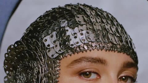 A close-up, profile-angled studio portrait of actress Florence Pugh. She wears an intricate, metallic helmet-style cap resembling overlapping metal chainmail scales, adorned with a large rosette emblem over the ear and a fine, spiky textured fringe framing her forehead and ears. Her expression is serene and contemplative, featuring soft, glowing makeup, a warm blush on her cheeks, and natural, well-defined brows. The background is a soft, muted grey with a curved dark blue architectural border hugging the lower left edge of the frame.
