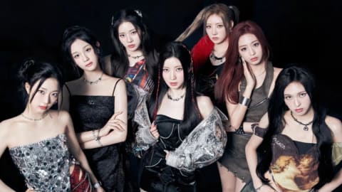 An group of seven young women pose together closely against a dark background, showcasing an edgy, modern fashion aesthetic. On the far left, a woman with dark hair tied back wears a strapless, glittering silver-and-black top paired with textured red trousers and a silver chain belt. Next to her, a woman in a black spaghetti-strap dress stands with her arms crossed, sporting a matching dark necklace. Behind them, a woman with long dark hair and a plaid-accented top looks forward. In the center, a woman with braided sections in her dark hair wears a black strapless dress under an oversized metallic silver jacket, paired with fishnet stockings. To her right, a woman with high pigtails and a dark top with a red fuzzy collar glances slightly sideways. Next to her, a woman with long red-toned hair poses with her hand to her cheek, dressed in an olive-green sleeveless top and matching skirt. On the far right, a woman with long, wavy dark hair wears a top featuring a classic art print of cherubs, paired with a pleated grey denim miniskirt. All members present a confident, synchronized expression toward the camera under cool-toned studio lighting.
