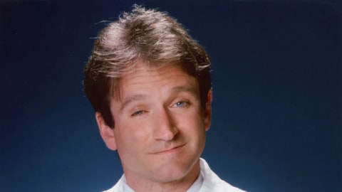 The image features a studio portrait of a young Robin Williams, captured from the chest up against a solid blue background. He is looking directly at the camera with a subtle, knowing smirk and slightly raised eyebrows, conveying his signature playful and charismatic energy. He is dressed in a stylish 1980s-style oversized white blazer with fine, light pinstripes, worn over a crisp white button-down shirt. A textured, light turquoise knit tie serves as a bright pop of color in the center of the frame. His arms are tightly crossed over his chest, and his dark brown hair is styled in a voluminous, feathered look characteristic of the era. The lighting is soft and even, highlighting his expressive facial features.