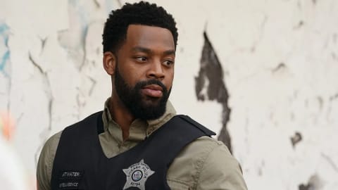 A medium shot of Officer Kevin Atwater, played by LaRoyce Hawkins, from the television series "Chicago P.D." He is standing outdoors against a weathered, peeling white wall, looking off to the side with a serious and focused expression. He wears an olive green button-down shirt under a black tactical police vest. The vest features a silver Chicago Police star badge with the number 52784 pinned to the chest, alongside nametags that read "ATWATER" and "INTELLIGENCE."