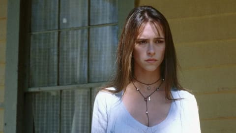 A medium shot captures a young woman with a concerned, somber expression, played by Jennifer Love Hewitt as Julie James in the 1997 horror film I Know What You Did Last Summer. She stands outdoors against a light-yellow wooden wall, with a large, multi-paned window featuring white lace curtains visible on the left. She has long, straight brown hair with wispy bangs framing her face, and is wearing a light blue or white knit top with a wide neckline. Layered around her neck are two thin necklaces: a shorter black cord choker with a small silver charm, and a longer delicate necklace adorned with small pearls and a hanging silver fish pendant that drops down her chest. The lighting is soft and natural, emphasizing her dark eyes and furrowed brow.