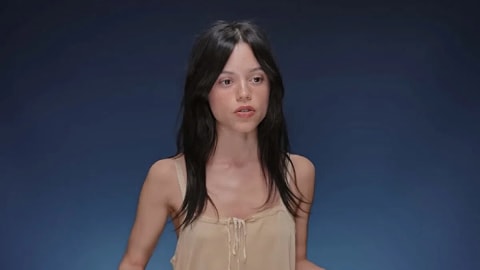 An over-the-shoulder medium shot features actress Jenna Ortega speaking directly to the camera against a solid navy blue studio backdrop. She has middle-parted, dark shoulder-length hair with soft curtain bangs framing her face. She wears a minimalist, beige sleeveless camisole top with delicate tie-front detailing and lace trim around the neckline. Her expression is focused and animated mid-speech, lit by soft, even studio lighting that highlights her features. An interview still shows actress Jenna Ortega from the waist up against a solid, dark blue gradient backdrop. She has dark, shoulder-length hair with soft curtain bangs framed around her face, and she is looking slightly to her right with an open mouth as if speaking. She wears a lightweight, sleeveless beige or light tan top featuring delicate thin straps and a small tie detail at the center of the scoop neckline.