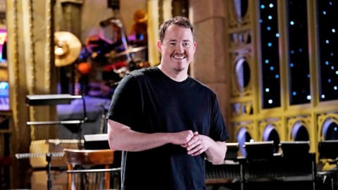 A man with short brown hair, a mustache, and a soul patch smiles widely while standing on a stage with his hands clasped together in front of him. He is wearing a plain black crewneck t-shirt and grey jeans. The stage behind him features musical instruments, microphone stands, an orchestral setup, and tall, gold-trimmed arched windows adorned with small blue lights that resemble a starry night sky.