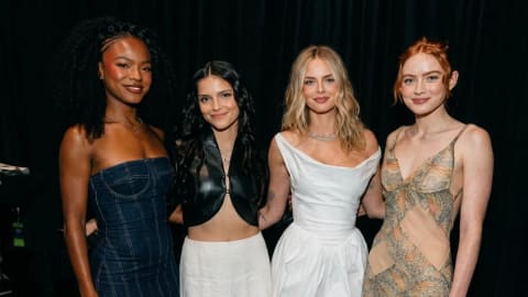 A medium-shot portrait captures Maya Boyd, Inde Navarrette, Samara Weaving, and Sadie Sink standing closely together against a dark, plain black backdrop. On the far left, Maya Boyd poses with a soft smile, styled with voluminous, dark curly hair pinned back at the sides. She wears a dark blue strapless denim maxi dress featuring visible pinstripes and structured seam lines along the bodice. Standing next to her, Inde Navarrette smiles directly at the camera with long, wavy dark hair framing her face. She wears an edgy black leather or faux-leather halter-neck crop top that splits open at the lower front, paired with a high-waisted, flowing white maxi skirt. Next in line, Samara Weaving smiles subtly, showcasing shoulder-length blond waves parted down the middle. She wears a structured, sleeveless white cocktail dress with a draped cowl neckline, a fitted waist, and a slight balloon silhouette at the skirt, complemented by a delicate silver necklace. On the far right, Sadie Sink smiles warmly with her signature red hair styled into an elegant updos with loose strands framing her face. She is dressed in an intricate, sleeveless V-neck maxi dress adorned with semi-sheer mesh paneling across the midriff and tiered, ruffled fabric detailing in muted tones of beige, green, and orange. The lighting is direct and bright, casting soft shadows behind the women and highlighting the varying textures of their outfits.