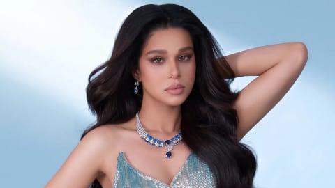A medium studio shot of Misbah Arshad posing gracefully against a soft blue gradient background. She is wearing a glamorous, strapless evening gown adorned with shimmering silver and teal sequins that form a patterned texture across the fitted bodice and ruched skirt. Her long, voluminous dark hair is styled in loose waves cascade over her shoulders, and one arm is raised behind her head. She accessorizes the elegant look with a matching silver and blue gemstone necklace and drop earrings, while gazing directly forward with a soft, neutral expression.
