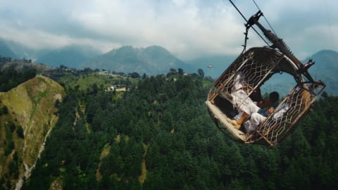 A wide aerial drone shot captures a makeshift, open-air metal cable car dangling dangerously at a sharp tilt high above a vast, densely forested mountain ravine. Inside the cramped basket structure, multiple passengers, including schoolboys in white shirts, are seen clinging tightly to the wire mesh frame. In the soft-focused background, rolling green hills and rugged mountain ridges rise under a vast, cloud-filled sky, emphasizing the extreme altitude and isolation of the stranded vehicle.