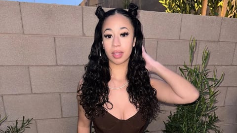 An eye-level, medium shot shows Jaden Newman standing outdoors against a light gray brick wall and a small green bush. She looks at the camera, smiling slightly. Her long, black, wavy hair is styled in two buns on top of her head with the rest cascading down her shoulders. Her face is made up with dramatic makeup, including dark eyeliner, pink lipstick, and dark eyeshadow. She wears a brown crop top, printed pants, and a thin silver necklace. The background is a gray brick wall with a green bush on the right side and dry leaves on the ground.