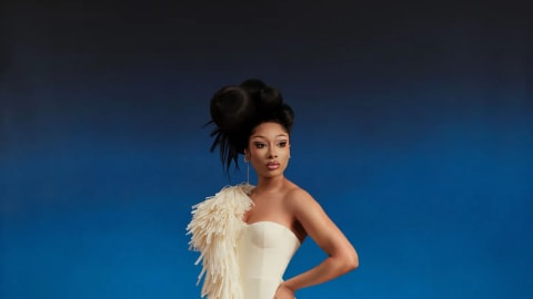Megan Thee Stallion strikes a powerful pose for InStyle's Fall 2026 Fashion and Beauty Issue. Captured against a dual-toned blue backdrop and standing amidst waves of vibrant red faux-fur, she wears a striking ivory Asymmetric Raffia Gown by designer Aiste Hong. The sculptural, strapless mermaid-silhouette dress features dramatic fringe details that wrap over her right shoulder and cascade down into a voluminous textured skirt. Complementing her avant-garde gown, her dark hair is styled into an intricate, structural updo with dynamic sweeping shapes, paired with refined drop earrings and a meticulous neutral glam look.