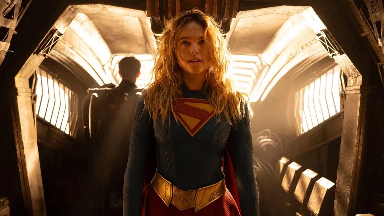 The image depicts Alcock's Supergirl in her superhero suit, featuring a dark blue textured material with a prominent red and gold "S" emblem on the chest. She has long, wavy blonde hair and wears a thick gold belt with a pointed buckle. She is standing within what appears to be a dimly lit, high-tech spaceship interior with warm, glowing lights.