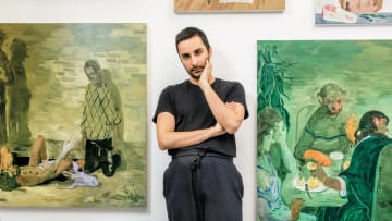 A medium shot showcases the Pakistani-American contemporary artist Salman Toor standing with a pensive expression in the center of his studio. He is dressed casually in a black crewneck T-shirt and dark gray sweatpants, with his left arm crossed tightly over his stomach and his right hand gently propping up his chin. Flanking him on either wall are two of his distinctive figurative oil paintings rendered in his signature, atmospheric emerald-green and ochre palettes. The artwork on the left depicts a surreal scene with a kneeling figure and another figure reclined on the ground, while the large canvas on the right illustrates an intimate, stylized gathering of multiple figures seated around a dinner table under warm lighting. A couple of smaller, unfinished canvases are mounted higher up on the white wall behind him.