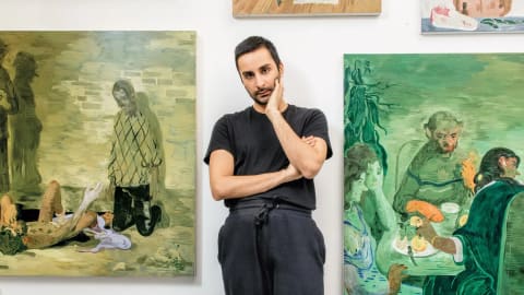 A medium shot showcases the Pakistani-American contemporary artist Salman Toor standing with a pensive expression in the center of his studio. He is dressed casually in a black crewneck T-shirt and dark gray sweatpants, with his left arm crossed tightly over his stomach and his right hand gently propping up his chin. Flanking him on either wall are two of his distinctive figurative oil paintings rendered in his signature, atmospheric emerald-green and ochre palettes. The artwork on the left depicts a surreal scene with a kneeling figure and another figure reclined on the ground, while the large canvas on the right illustrates an intimate, stylized gathering of multiple figures seated around a dinner table under warm lighting. A couple of smaller, unfinished canvases are mounted higher up on the white wall behind him.