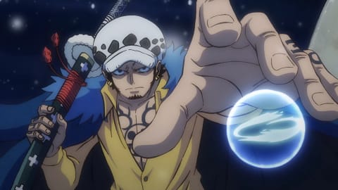 An anime screenshot features Trafalgar Law from One Piece, looking forward with a serious, determined expression against a dark background with hints of light. He wears his signature white and black spotted hat, a yellow button-up shirt left open at the collar to reveal his intricate black chest tattoos, and a dark blue coat lined with fur around the shoulders. In his right hand, he holds the hilt of his large sword, the Kikoku, which is wrapped with red cord and decorated with light cross-shaped symbols. His left hand is thrust prominently toward the viewer in the foreground, fingers slightly curled, with a small, glowing blue energy sphere floating just beneath his palm, symbolizing his "Room" ability.