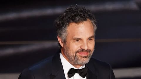An older, middle-aged man with salt-and-pepper hair and a matching short beard smiles gently while posing for a photograph. He is wearing a formal black tuxedo jacket with sleek satin lapels over a crisp white button-down dress shirt. A classic black bowtie is neatly fastened at his collar. His dark curly hair is styled naturally with some volume, and his warm brown eyes look slightly off-camera. The background is a softly blurred, dark, textured surface with horizontal lines, suggesting an indoor red carpet or awards ceremony venue.