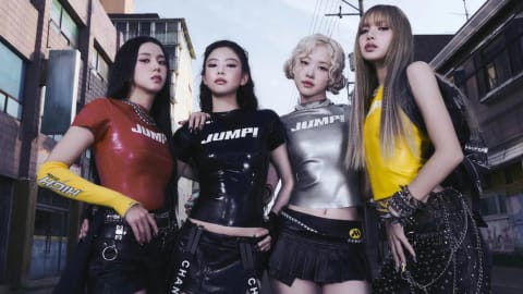 BLACKPINK stands firmly side-by-side in a gritty, industrial street setting under an open sky, radiating a powerful and modern aesthetic. The members pose in highly coordinated, edgy streetwear outfits featuring sleek, tight-fitting tops emblazoned with bold athletic branding. Jisoo commands attention on the left in a vibrant red latex shirt paired with a distinctive yellow arm sleeve. Next to her, Jennie wears a gloss-black "JUMPI" t-shirt over a structured, branded belt skirt. Rosé shines with pale blonde curls in a metallic silver mock-neck top and a micro pleated skirt, while Lisa anchors the right side of the frame in a bright yellow sleeveless top, sporting her signature long bangs and intricate heavy-metal chain detailing across dark pants.