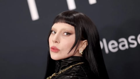 Lady Gaga delivers a striking visual statement on the red carpet with a high-drama beauty look defined by ultra-short micro bangs, long raven-black hair, and completely bleached eyebrows. Her complexion is flawless and luminous, paired with a sharp, subtle winged eyeliner that draws focus to her eyes, and a soft, matte rose lip color. She wears a high-necked, avant-garde black textured garment featuring intricate lacing details, accessorized with a statement gold chain necklace and delicate, light-colored earrings that catch the light, all set against a dark backdrop with blurred event signage.