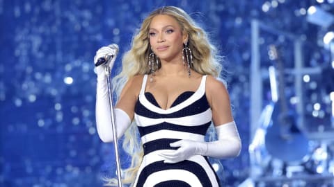 Beyoncé commands the stage during her Renaissance World Tour performance, radiating elegance in a custom black-and-white geometric gown. The striking ensemble features a deep sweetheart neckline and an intricate pattern of swirling optical lines that accentuate her silhouette. Complementing the bold dress are pristine white opera-length gloves and dazzling, tiered crystal drop earrings that shimmer under the stadium lights. With her voluminous blonde hair styled in effortless, cascading waves, she holds a silver microphone against a glittering, blue-toned backdrop of reflective sequins and stage rigging, epitomizing high-fashion concert glamour.