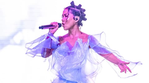 An elegant live stage performance showcasing a fluid, sculptural outfit and creative hairstyling. The singer Jhené Aiko performs onstage in a striking, ethereal lilac gown during a live musical concert. The dress features delicate, sheer organza overlays that flow dynamically around her arms and bodice, creating a fluid, sculptural silhouette illuminated by dramatic stage lighting. Her hair is styled in structured, artistic Bantu knots, and she holds a microphone to her lips while wearing custom in-ear monitors, delivering an expressive, high-energy vocal performance against a bright, minimalist backdrop.