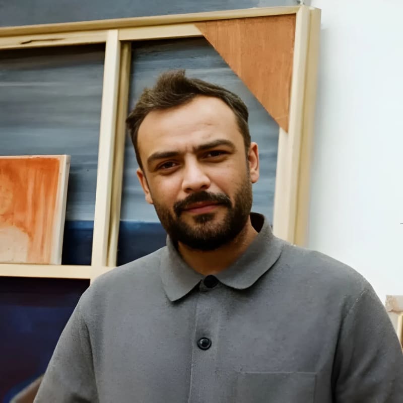 An average-looking adult man (Suleman Aqeel Khilji) with tanned skin, a neat dark beard, and short hair stands in a brightly lit art studio, looking directly at the camera with a gentle, relaxed expression. He is wearing a grey buttoned long-sleeve shirt jacket with a pocket on the left breast and dark grey pants. He is standing in the middle of the frame, surrounded by large, unframed paintings. Behind him on the left, a large canvas stretcher leans against the wall, revealing smaller rectangular canvas paintings peeking out. The surrounding artwork features earthy tones of brown, tan, and blue, depicting abstract landscapes and figures. The studio has a light-colored floor and white walls.