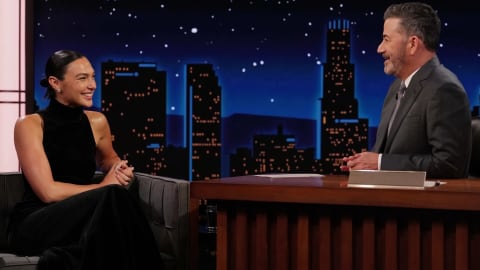 Late-night laughs with Wonder Woman. An evening talk show interview featuring Jimmy Kimmel conversing with actress Gal Gadot on the set of Jimmy Kimmel Live!, set against a stylized nighttime city skyline background. Gal Gadot sits comfortably on a grey chair wearing a sleek, black halter-top dress while smiling warmly toward Jimmy Kimmel, who sits across from her at his signature wooden desk wearing a sharp grey suit and tie. A large lower-third graphic overlays the bottom of the screen displaying the show's logo alongside Gal Gadot's name in bold white text.