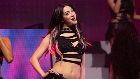 The image captures American singer and dancer Megan Skiendiel performing dynamically on stage, her dark hair accented with vibrant pink highlights that catch the theatrical lighting. She is dressed in an edgy, modern performance outfit featuring a black multi-cutout crop top revealing a sequined bralette underneath, paired with a studded choker necklace that enhances the rebellious, pop-rock aesthetic of the production. Her expression is animated and engaging as she commands the spotlight during a live musical set, while background elements hint at the energetic atmosphere of a full concert production with accompanying dancers and LED screens.