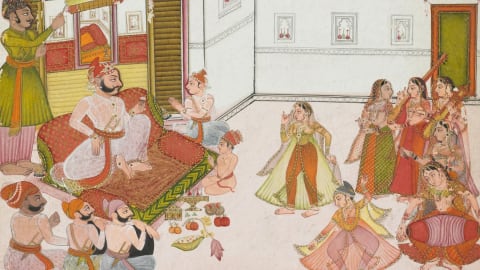 An opaque watercolor and ink painting on paper depicts a lively courtly gathering within a white-walled terrace or open courtyard. On the left, a bearded ruler with a prominent mustache sits cross-legged on a large, low, patterned cushion supported by red bolsters. He is dressed in a translucent white jama robe, an ornate red and gold turban, and fine jewelry as he looks toward a performance. Behind him, a standing attendant in a green gown holds a fly-whisk, while in front of his platform, two young boys sit on the floor, one with hands pressed together in a respectful gesture. In the lower-left corner, three male courtiers in turbans sit on the ground with their backs partially turned to the viewer.The right side of the painting centers on the entertainment, where two female dancers perform; one stands elegantly in a yellow and green dress, while another spins dynamically in a pink outfit. They are accompanied by a tight cluster of five female musicians on the far right playing traditional instruments, including a stringed tanpura and a small dholak hand drum. The architectural setting features a white wall adorned with small rectangular niches containing red decorative vessels, alongside an open green doorway on the far right. In the foreground between the ruler and the performers, a careful arrangement of offerings is spread across the floor, featuring small golden containers, a lotus bud, and fresh fruits like oranges and pomegranates.