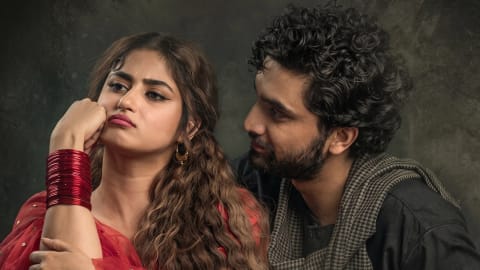 A close-up studio promotional photograph captures Pakistani actors Sajal Aly and Ahad Raza Mir in character from the period drama serial Aangan. Sajal Aly, portraying the character Chammi, sits on the left side of the frame looking away to the left with a highly expressive, visibly annoyed, and pouting facial expression. Her right arm is raised with her fist supporting her chin, showcasing thick, metallic red glass bangles covering her wrist. She wears traditional South Asian attire consisting of a red and gold embroidered dress paired with a sheer red georgette dupatta draped over her shoulders. Her long, dark brown hair is styled in voluminous, crimped waves cascading down her front, complemented by bold winged eyeliner, defined eyebrows, and matte mauve lipstick. To her right, Ahad Raza Mir, portraying the character Jamil, leans in closely from behind her right shoulder, gazing at her with a gentle, micro-smiling, and placating expression. He features thick, curly black hair, a neatly trimmed beard, and is dressed in a dark grey kurta layered with a grey-and-black micro-check pattern scarf loosely draped around his neck. The portrait is set against a dark, textured studio background with mottled grey and charcoal tones, evoking a dramatic, vintage atmosphere.