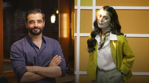 A side-by-side split-screen image features Pakistani actors Hamza Ali Abbasi and Hania Amir. On the left side, Hamza Ali Abbasi stands with his arms crossed over his chest, facing forward with a gentle smile. He has short, slicked-back dark hair, a neatly trimmed beard, and a mustache. He is wearing a dark, charcoal-gray, long-sleeved button-up shirt with the collar slightly open. The background behind him is softly blurred, showing a warm, ambient-lit indoor setting with horizontal window blinds and a glowing light fixture. On the right side, Hania Amir stands angled slightly to the side while looking toward the camera with a subtle expression. Her long, dark, wavy hair is parted down the middle, with a portion pulled back. She wears a white, wide-collared button-down shirt beneath a bright, lime-yellow tailored blazer and light sage-green trousers with her left hand tucked into the pocket. An elegant, patterned silk scarf in shades of olive, white, and yellow is tied around her neck, and she wears silver hoop earrings. The background behind her is a prominent geometric grid design with yellow and cream-colored rectangular panels. Edited by PakistaniArt.
