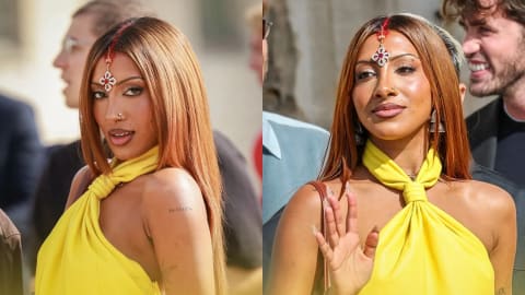 A side-by-side composite photo shows Lara Raj of the girl group KATSEYE outdoors, looking toward the camera. In both frames, she has long, straight, copper-brown hair parted down the middle, featuring a streak of red color along the part line and a decorative jeweled maang tikka forehead ornament. She is wearing a vibrant yellow halter-neck top or dress that twists at the front of the neck. In the left frame, she casts a sharp glance over her shoulder, showing a small text tattoo on her upper arm, while in the right frame, she waves one hand with neutral makeup and a nose ring visible.