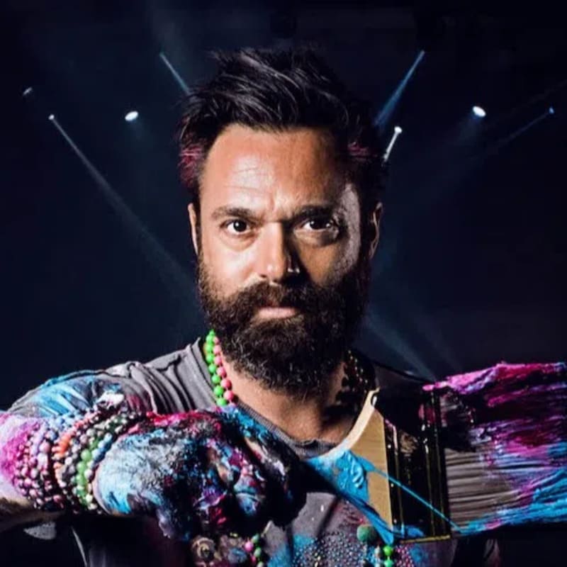 British contemporary artist Sacha Jafri is pictured covered in vibrant paint splatters while holding a large brush used to create his monumental masterpiece, The Journey of Humanity. Created over seven months during the 2020 lockdown inside the ballroom of Atlantis, The Palm in Dubai, this colossal record-breaking creation spans over 17,000 square feet—making it officially recognized by [Guinness World Records](1.2.1, 1.3.2) as the largest art canvas in the world. The sprawling abstract work aims to foster global connectivity and empathy, incorporating artwork contributed online by children from more than 140 countries. The entire canvas was ultimately split into 70 framed sections and sold at a humanitarian auction for a staggering $62 million, with all proceeds dedicated to children's educational and health charities globally.