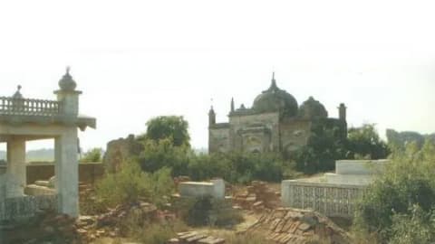 A landscape photograph showcases the historic Miani Mosque in Hyderabad, Sindh, Pakistan, an ancient brick structure featuring a prominent central dome and arched entryways that sits in a state of partial dilapidation amid dense wild vegetation. In the foreground, the crumbling ruins of brick walls and weathered stone graves litter the overgrown ground, leading towards a modern white, intricately carved balustrade and pavilion structure on the left side of the frame. The sky above is entirely blown out and bright white, casting a harsh, flat light over the historic site and its surrounding burial grounds.