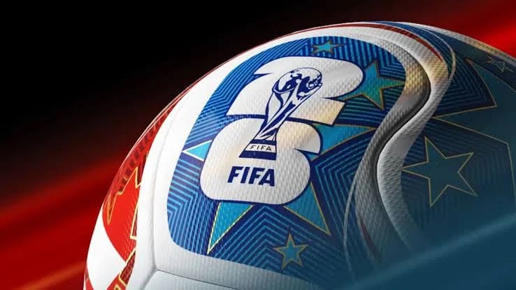 An extreme close-up shot captures the official match ball for the FIFA World Cup 2026™, angled from a low perspective against a dynamic background of blurred red and black streaks. The curved surface of the white soccer ball features intricate, modern textures composed of fine micro-grooves and debossed patterns that trace the contours of its thermally bonded panels. Prominently displayed on one section is the stylized "26" tournament emblem, rendered in white with a bold blue border. Superimposed directly over the center of the numeral is a sharp, detailed graphic of the FIFA World Cup Trophy, with the word "FIFA" printed clearly in a crisp, dark blue sans-serif font beneath it. The surrounding graphic elements include a vibrant pattern of turquoise and deep blue fields overlaid with golden-outlined, five-pointed teal stars, all converging to create a sleek, high-tech, and futuristic sports aesthetic.