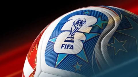 An extreme close-up shot captures the official match ball for the FIFA World Cup 2026™, angled from a low perspective against a dynamic background of blurred red and black streaks. The curved surface of the white soccer ball features intricate, modern textures composed of fine micro-grooves and debossed patterns that trace the contours of its thermally bonded panels. Prominently displayed on one section is the stylized "26" tournament emblem, rendered in white with a bold blue border. Superimposed directly over the center of the numeral is a sharp, detailed graphic of the FIFA World Cup Trophy, with the word "FIFA" printed clearly in a crisp, dark blue sans-serif font beneath it. The surrounding graphic elements include a vibrant pattern of turquoise and deep blue fields overlaid with golden-outlined, five-pointed teal stars, all converging to create a sleek, high-tech, and futuristic sports aesthetic.