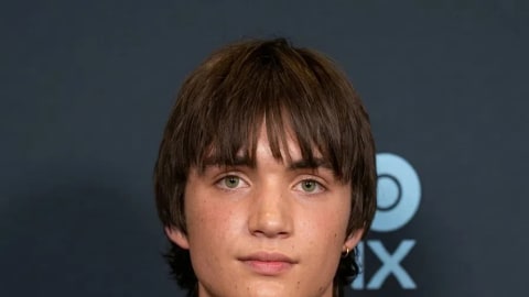 The image features actor Aran Murphy, son of Cillian Murphy, posing at a red carpet event against a dark blue background adorned with a partial "DX" logo. He is looking directly at the camera with a subtle, neutral expression, sporting a textured brown bowl-cut hairstyle with long bangs framing his eyes, and a hint of a small gold hoop earring in his left ear. He is wearing a cozy, thick grey wool knit sweater featuring an open, slightly distressed texture with visible white spots or under-layer accents peeking through, worn over a simple white crew-neck undershirt and a thin gold chain necklace.