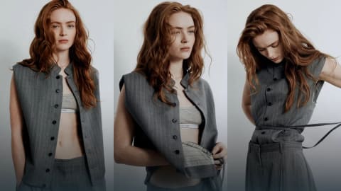 Sadie Sink presents an effortless fusion of structured tailoring and modern streetwear, capturing a relaxed yet highly polished aesthetic. Dressed in an ensemble from Calvin Klein's Spring 2027 Collection designed by Veronica Leoni, she pairs an elongated, high-neck sleeveless pinstripe vest featuring dark button details with matching relaxed-fit pinstripe trousers. Underneath the vest, a classic grey cotton Calvin Klein bralette peeks through, introducing a sporty element to the formal suiting backdrop. The three-panel composition showcases her from different angles, highlighting the structural versatility of the outfit—from its loose, open drape to a tightly belted, defined waist profile styled with a slim coordinating wrap cord. Her vibrant, copper-toned hair falls in undone, textured waves over her shoulders, softening the sharp, corporate lines of the monochrome charcoal suit.