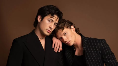 A formal portrait captures actors Hudson Williams and Connor Storrie posing closely against a solid brown studio backdrop. On the left, Williams stands facing slightly right, wearing a sleek, double-breasted black suit jacket with wide lapels and matching trousers, accented by a subtle silver chain necklace. Storrie stands to his right, gently resting his head against Williams’s left shoulder while looking directly forward with a soft, serious expression. He wears a dark, pinstriped suit jacket left unbuttoned over a black scoop-neck tank top, paired with matching pinstriped trousers. The lighting creates a high-contrast, moody atmosphere, highlighting their expressions and the tailoring of their outfits.