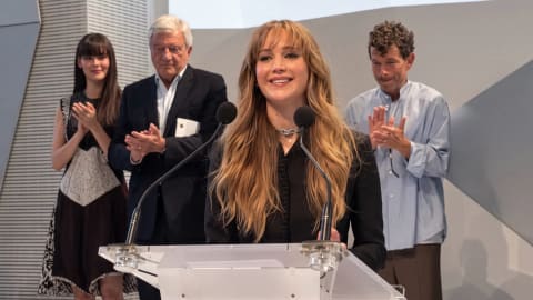An eye-level medium shot captures American actress Jennifer Lawrence speaking smilingly from behind a transparent acrylic podium equipped with dual microphones. She has long wavy blonde hair with bangs and wears a tailored black jacket and a metallic choker. Standing in the background and applauding her are three individuals: a young woman on the left in a patterned dress, an older gentleman in a dark suit jacket, and a man on the right wearing a loose light blue button-down shirt and brown trousers. The setting features a modern, minimalist white stage with sharp geometric architectural angles.