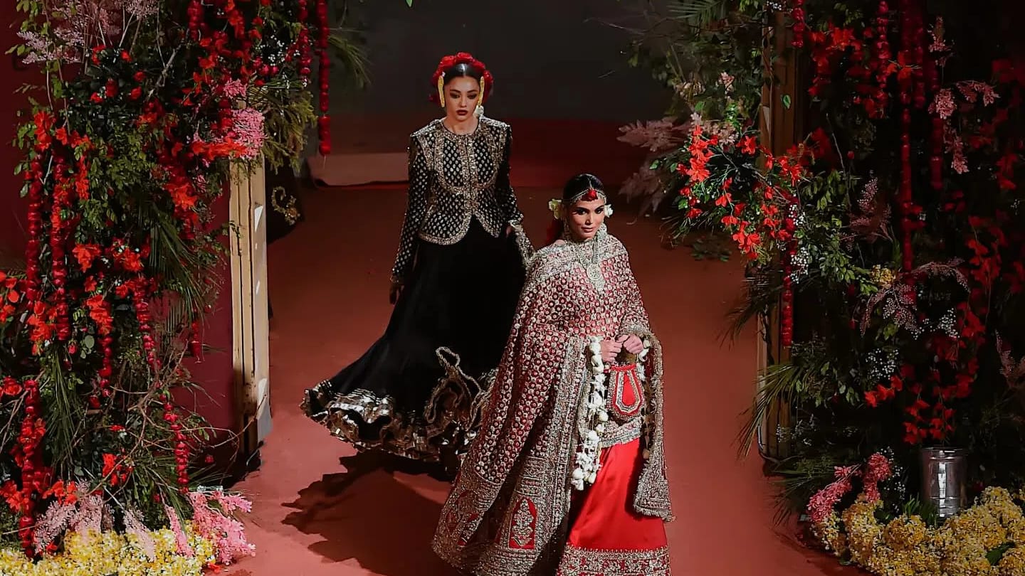 A wide, high-angle shot captures two models presenting the "Nargis" Bridal Couture SS 26 collection on an indoor runway floored with solid terracotta-red carpeting.Foreground Model: Walking confidently forward on the right, she wears a heavily traditional red bridal lehenga. The ensemble features a flowing crimson skirt hemmed with thick gold embroidery and a matching, heavily draped shawl-style dupatta covering her torso and arms. Her styling includes a traditional maang tikka, dangling earrings, a sleek middle-parted low bun, and an elaborate, dangling white-flower kiran garland clutched alongside a small bag.Background Model: Walking slightly behind and to the left, she models an elegant jet-black lehenga choli. The outfit comprises a structural, full-sleeved black jacket-style choli covered in geometric gold zari grids and a wide, layered black skirt that flares dramatically toward the hem with matching gold trimmings. A crown of bright red roses accents her slicked-back hairstyle.Framing the runway are massive, asymmetrical floral installations. Dense arrangements of lush green foliage, trailing vines, and blooming crimson and soft-pink roses grow along the sides. Scattered yellow and white flower petals blanket the base of the arrangements.