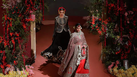 A wide, high-angle shot captures two models presenting the "Nargis" Bridal Couture SS 26 collection on an indoor runway floored with solid terracotta-red carpeting.Foreground Model: Walking confidently forward on the right, she wears a heavily traditional red bridal lehenga. The ensemble features a flowing crimson skirt hemmed with thick gold embroidery and a matching, heavily draped shawl-style dupatta covering her torso and arms. Her styling includes a traditional maang tikka, dangling earrings, a sleek middle-parted low bun, and an elaborate, dangling white-flower kiran garland clutched alongside a small bag.Background Model: Walking slightly behind and to the left, she models an elegant jet-black lehenga choli. The outfit comprises a structural, full-sleeved black jacket-style choli covered in geometric gold zari grids and a wide, layered black skirt that flares dramatically toward the hem with matching gold trimmings. A crown of bright red roses accents her slicked-back hairstyle.Framing the runway are massive, asymmetrical floral installations. Dense arrangements of lush green foliage, trailing vines, and blooming crimson and soft-pink roses grow along the sides. Scattered yellow and white flower petals blanket the base of the arrangements.