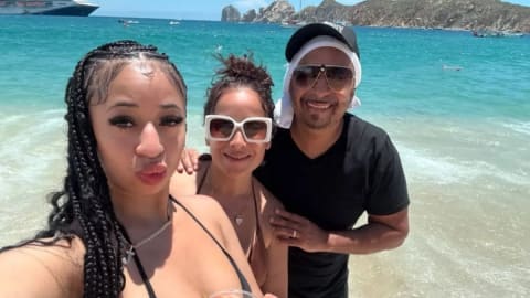 A sunny, eye-level selfie captures three people smiling together on a tropical beach with turquoise water and rocky cliffs in the background. In the foreground, a young woman with long black braids and a black bikini top holds a clear plastic cup filled with a yellow drink, leaning into the frame. Standing close behind her is a woman with her hair up, wearing large white-rimmed sunglasses and a neutral-toned swimsuit, who smiles warmly toward the camera. To her right, a man in a black T-shirt, aviator sunglasses, and a black baseball cap worn over a white towel or fabric smiles broadly, completing the group. The ocean water fades from a vibrant turquoise to a frothy white shore around their ankles, while a large cruise ship and small boats are visible in the distance under a clear blue sky.
