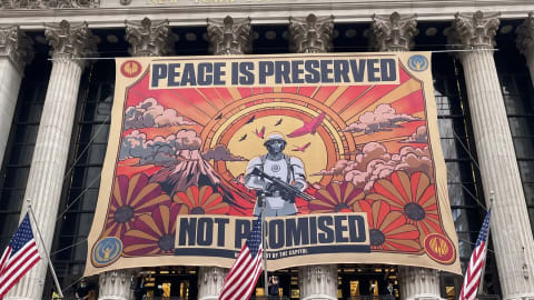 A massive, retro-style propaganda banner hangs across the classical columned facade of the New York Stock Exchange. The banner features a central soldier in white futuristic armor holding a rifle, set against a stylized landscape with a volcano, birds, clouds, and large sun rays radiating from behind him. Bright orange, pink, and yellow hues dominate the graphic, which is framed by red borders featuring circular logos. The bold text across the banner reads, "PEACE IS PRESERVED" at the top and "NOT PROMISED" across the bottom, with a smaller line below stating "VERIFIED BY THE CAPITOL." Three American flags are mounted on the building's balcony directly beneath the banner.