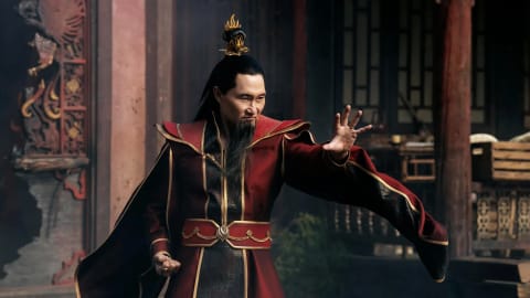 In this promotional still from the live-action adaptation of Avatar: The Last Airbender, actor Daniel Dae Kim perfectly embodies the tyrannical and ruthlessly driven ruler of the Fire Nation, Fire Lord Ozai. Clad in his highly detailed, regal crimson-and-black imperial robes accented with ornate gold trim, Ozai strikes an aggressive, commanding firebending stance with an outstretched open palm. His long black hair is partially pulled back into a traditional topknot secured by a sharp, flame-styled gold hairpiece, complementing his distinctive long, sharp goatee. The intense, calculating glare across his face highlights his absolute desire for total world conquest, captured in a dimly lit, traditional East Asian architectural courtyard that echoes the oppressive, industrial aesthetic of the Fire Nation Palace.