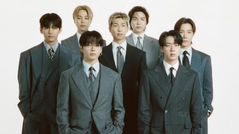 A group of seven young men, the globally acclaimed South Korean boy band BTS, pose together against a minimalist white studio background for a formal promotional poster. The members—Jungkook, Suga, J-Hope, RM, Jimin, V, and Jin—are arranged on stacked white rectangular platforms, dressed in sophisticated, monochromatic suits ranging from charcoal grey to deep black, paired with collared shirts and ties. Above them, a solid black banner prominently displays the text "ARTIST OF THE YEAR" in bold yellow lettering, while the bottom of the poster features a dark, cosmic outer-space graphic with the group's name "BTS" in large white font alongside official corporate logos for the MAMA Awards and Visa.