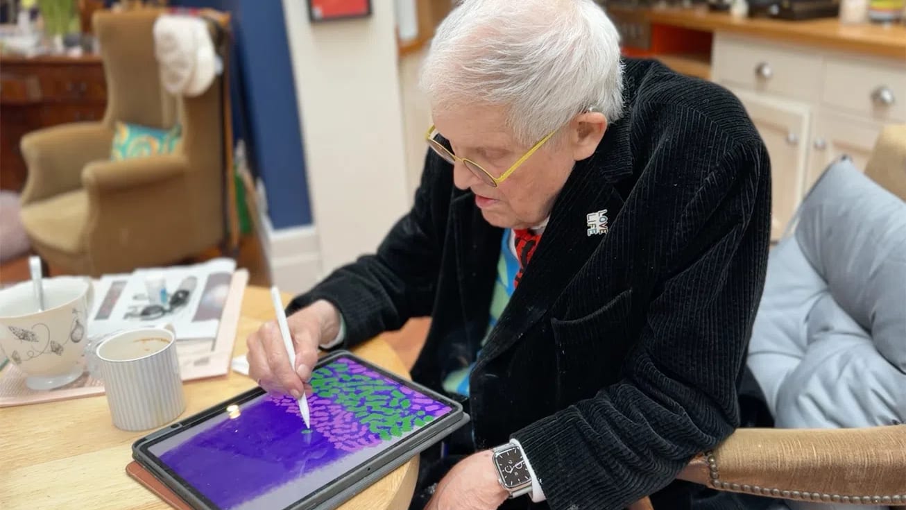 A close-up, slightly high-angle shot captures the influential British artist David Hockney seated at a light-colored wooden table, intently focused on creating digital art. Hockney, an elderly man with short white hair and yellow-rimmed eyeglasses, leans forward while wearing a black corduroy jacket adorned with a small "LOVE LIFE" pin on the lapel, layered over a colorful shirt and a patterned red tie. In his right hand, he holds a white stylus, carefully drawing vibrant green and pink brushstroke patterns across the glowing, deep purple screen of an iPad propped up on the table in front of him. On his left wrist, a distinctive square-faced wristwatch with a visible skeleton dial is prominent as his hand rests near his lap. The warm, softly blurred indoor background reveals a cozy studio or living space, featuring a tan armchair with a white hat resting on its back, a grey cushioned chair to his right, and a nearby table holding a white coffee mug and a patterned teacup.