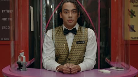A close-up shot from a music video featuring a man with curly hair and jewelry looking directly forward from behind a glass ticket booth. He is wearing a white dress shirt, a black bowtie, a green-and-gold patterned vest, and a rectangular name tag that reads "KEVIN". His hands are clasped in front of him on a pink curved counter surface. Outside the booth, posters and notices are visible, including one on the right that reads "NO INANIMATE OBJECTS." Yellow subtitle text at the bottom of the screen reads, "Sir, please leave." Dancer Kevin Scheitzbach appears as Kevin the Movie Ticket Guy in Taylor Swift's retro-styled "Opalite" music video.