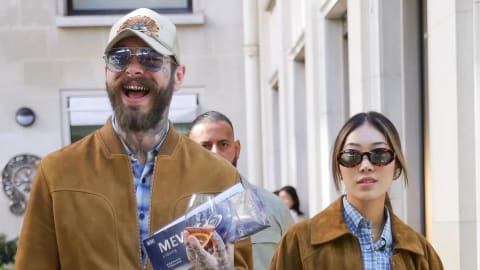 An image shows American musician Post Malone laughing while walking outdoors alongside his partner, Christy Lee, both wearing matching brown suede jackets over blue patterned tops. Post Malone, on the left, is wearing a structured brown suede bomber jacket over a blue and white checkered shirt, paired with light-wash blue jeans, a light-colored baseball cap, and aviator sunglasses, while holding a wine glass and a blue box of Mevius Lights cigarettes. Christy Lee, on the right, wears a loose-fitting brown suede jacket over a blue plaid mini dress secured with a brown leather belt, styled with tortoiseshell sunglasses and tall brown leather boots, as they walk down a paved pathway flanked by light-colored building facades with a security guard following closely behind. Post Malone and Christy Lee walk together in coordinating suede outfits. Photo by Backgrid.