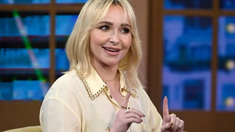 A medium shot captures American actress Hayden Panettiere from the chest up during a television talk show appearance. She is smiling cheerfully, facing slightly to her right while gesturing with her hands. Her blonde hair is styled in a shoulder-length bob with wispy curtain bangs. She wears a cream-colored, button-down blouse featuring a distinct gold chain-link trim along the edges of the collar. Her accessories include a nose stud, a delicate necklace, and rings on her fingers. The background features a softly blurred studio set with window panes overlooking a blue-toned city backdrop at night.